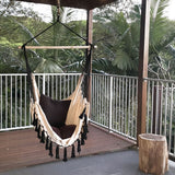 white hammock with black tassels hanging under a balcony in a lush garden next to a tree stump table