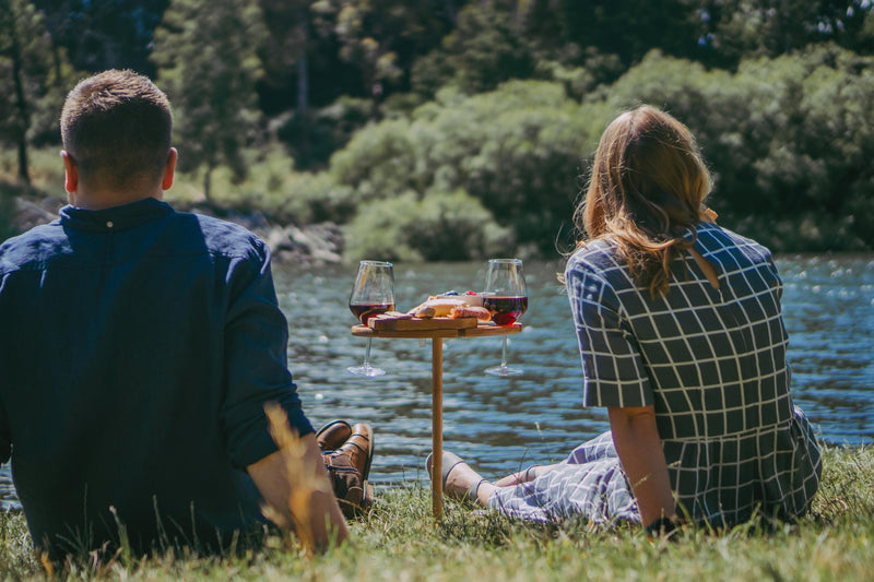 couple sitting by the river enjoying a wine and cheese platter on a bamboo picnic table